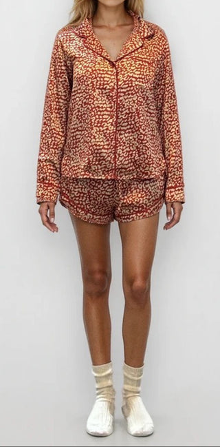 Woman wearing a red and gold leopard print pajama shorts and matching long sleeve button up pajama top. Plain white/grey backgorund.